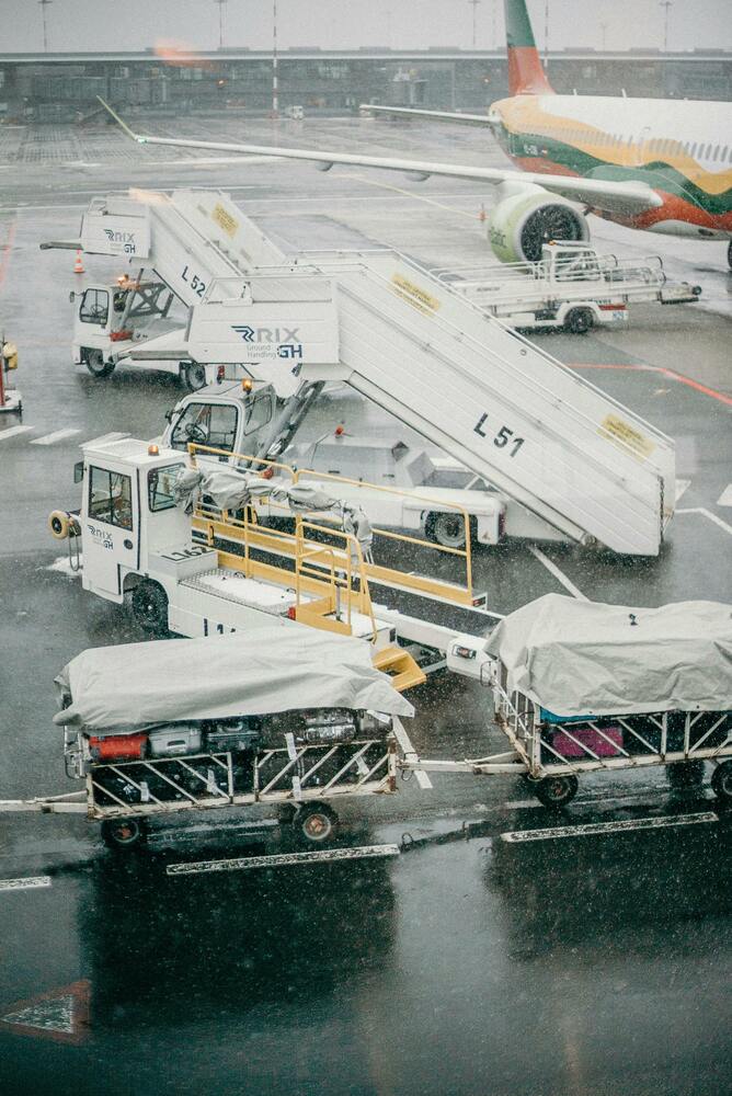 Quality pre-owned GSE, like luggage carts and stair vehicles, sit on a rainy airport tarmac near a parked airplane with a colorful tail.