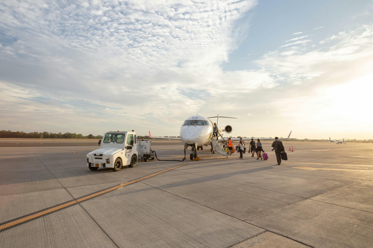 Passengers walk towards a small airplane on the tarmac, with a towing vehicle nearby under a bright sky.