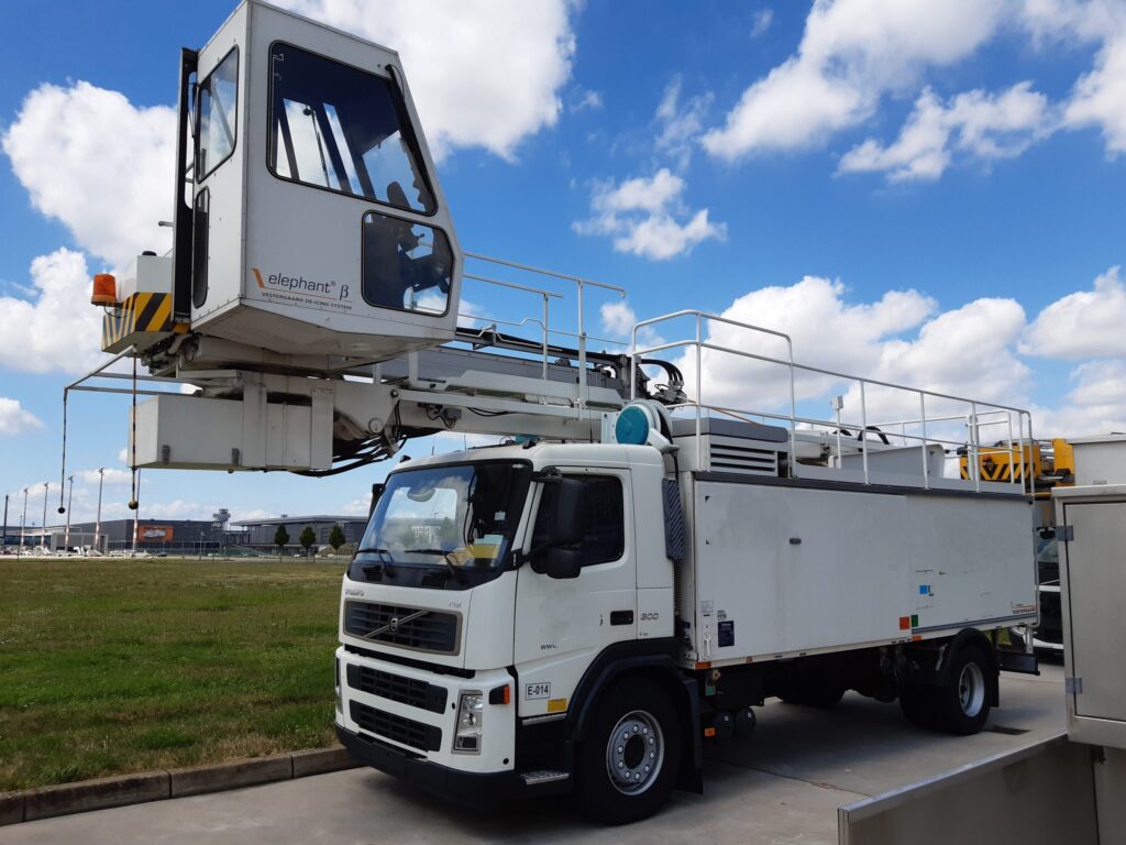 A white de-icing truck with an elevated cabin parked on a concrete surface near a grassy area, under a partly cloudy sky.