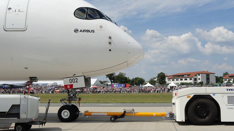 Close-up of an Airbus aircraft nose being towed on an airport tarmac with a crowd and buildings in the background.