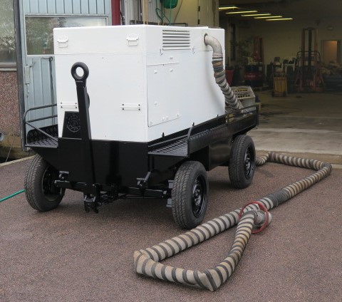 A white and black industrial machine with wheels, connected to large flexible hoses, is positioned outside a garage.