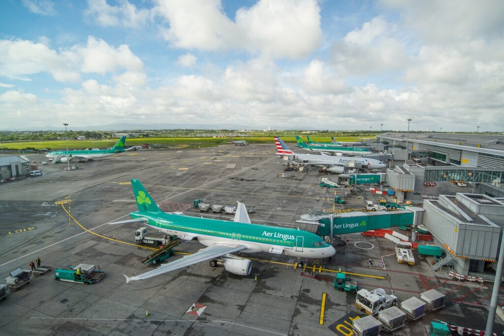 View of an airport terminal with several Aer Lingus aircraft parked at gates under a cloudy sky. Baggage carts and ground service vehicles are visible.