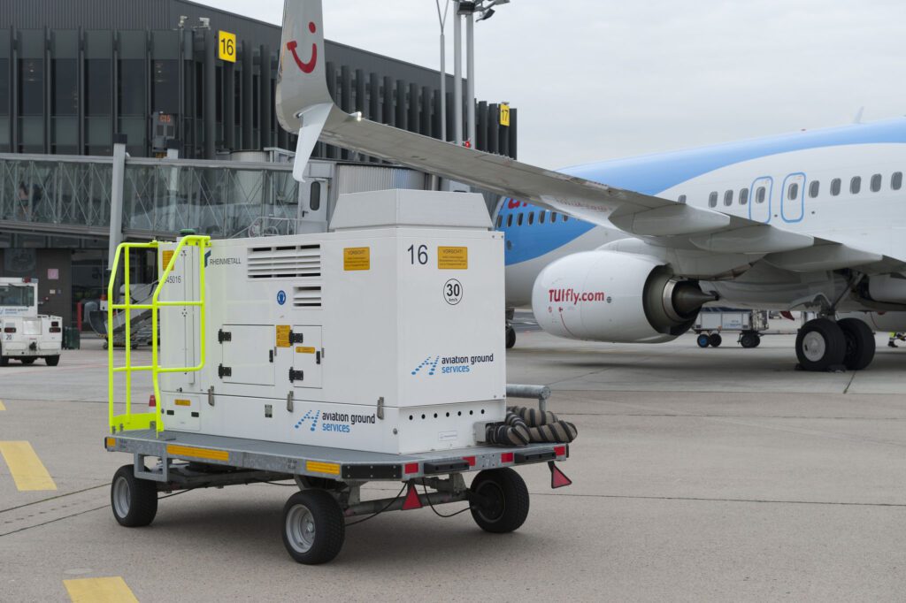 Ground power unit next to an airplane on airport tarmac, with terminal building in the background.