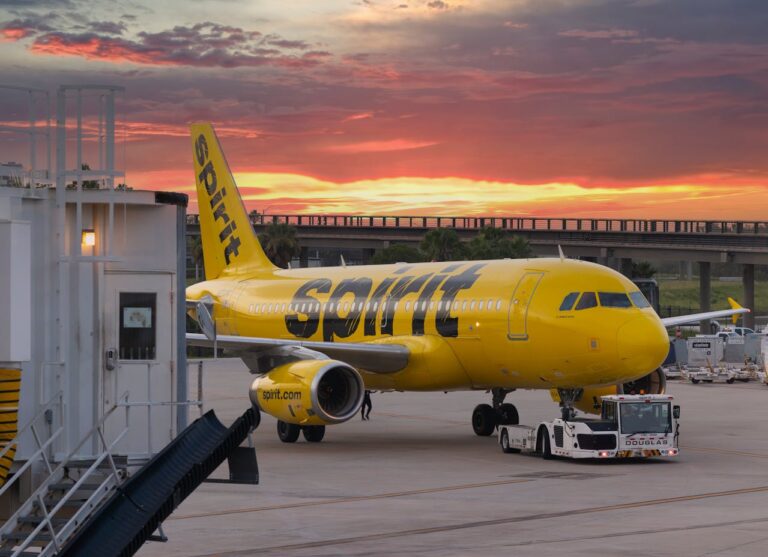 A Spirit Airlines plane is positioned on the tarmac at sunrise, with a tug vehicle in front and an airport gate nearby.