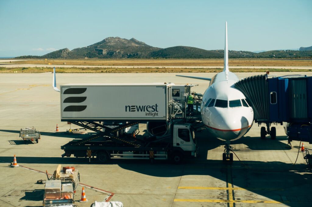 An airplane at an airport gate with a catering truck parked beside it. A jet bridge is connected, and mountains are visible in the background.