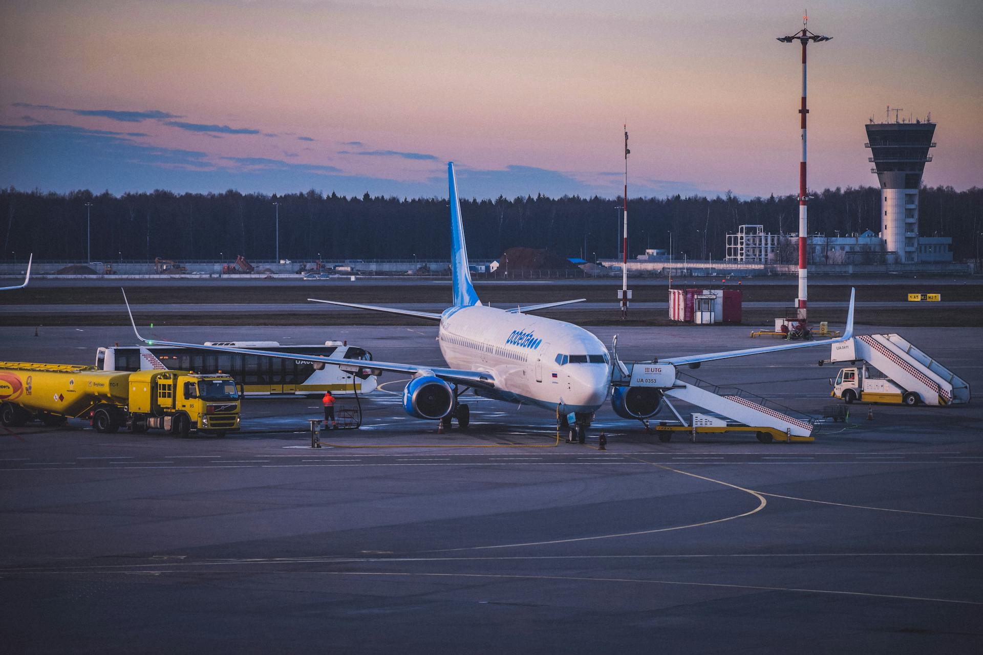 An airplane is parked at an airport, being serviced by ground crew with vehicles nearby. A control tower and a forest are visible in the background during sunset.