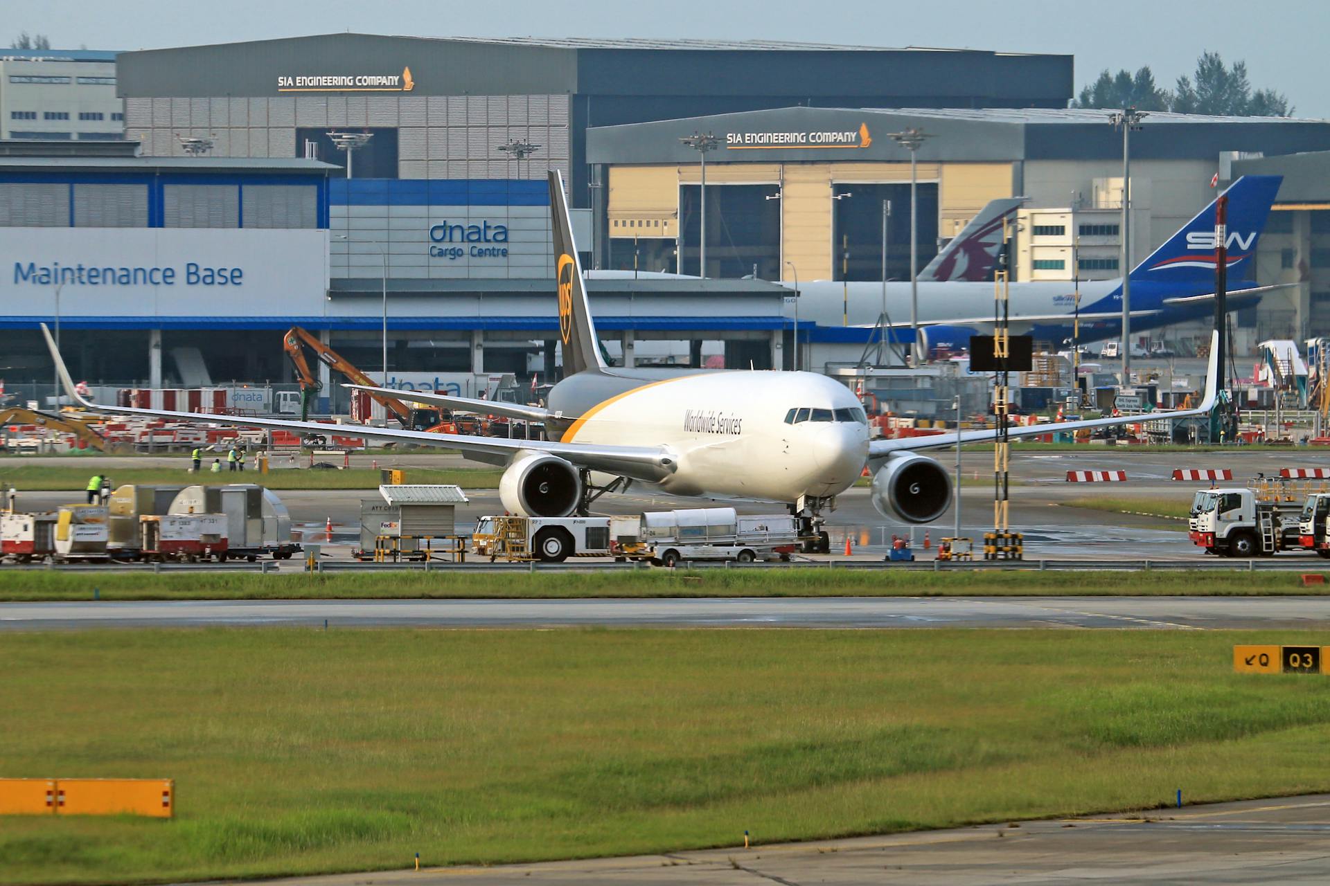 A large cargo plane is parked on the runway at an airport, with maintenance buildings and other aircraft in the background.