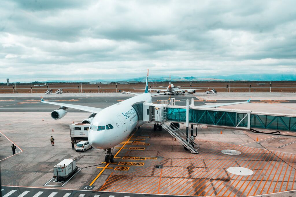 Airplane at an airport gate with a jet bridge attached, surrounded by vehicles and distant planes under a cloudy sky.