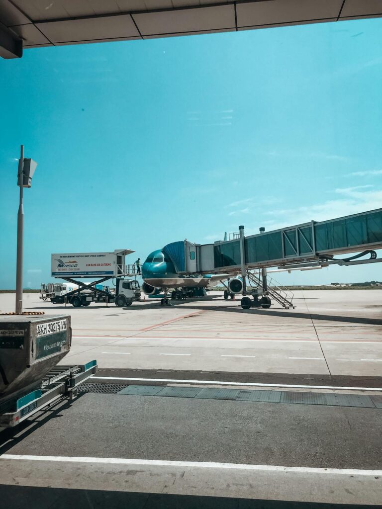 An airplane is parked at an airport gate while connected to a jet bridge. A catering truck is positioned next to the aircraft. The sky is clear and bright.