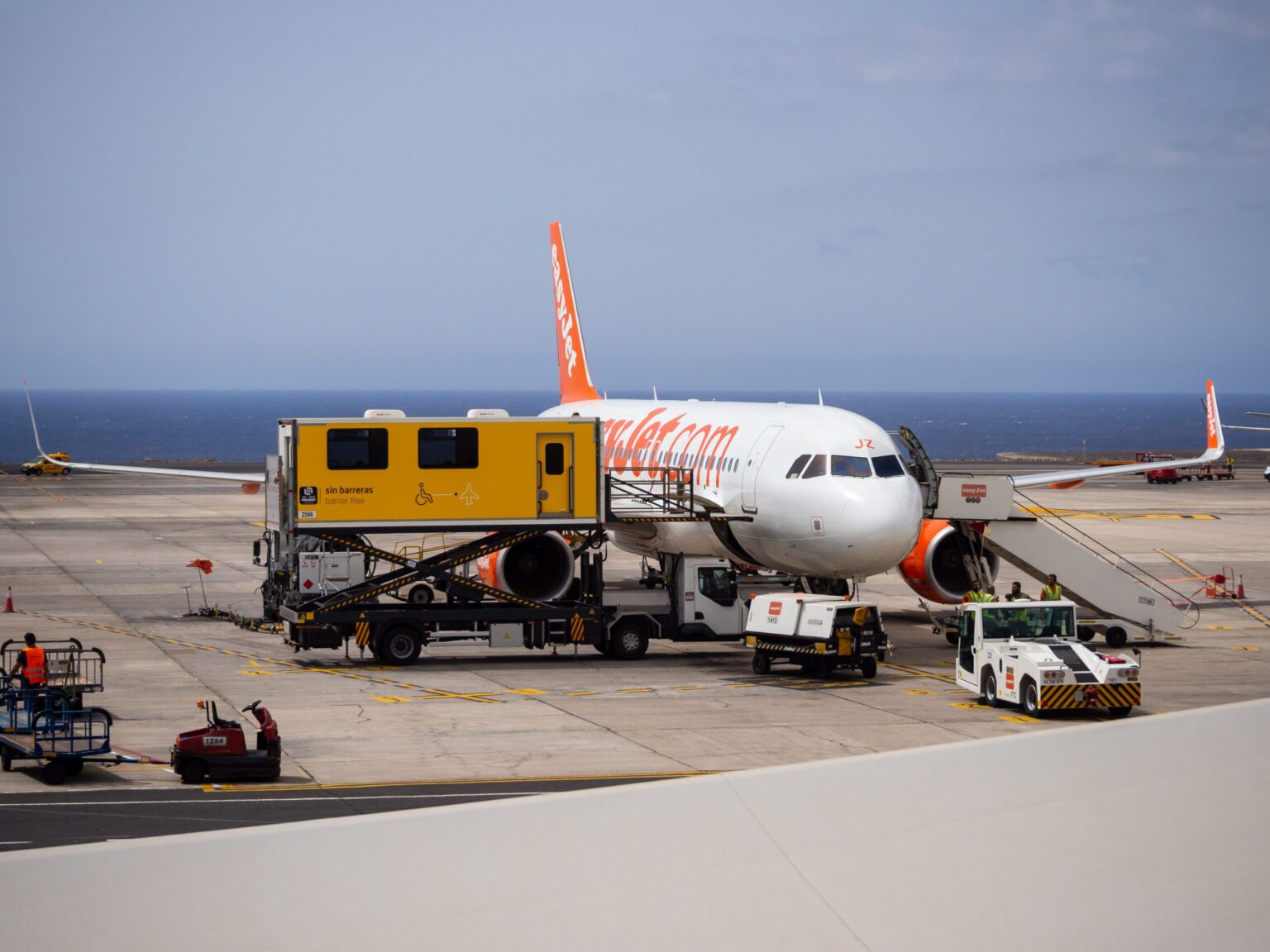An airplane is parked at the airport gate with a catering truck servicing it. Ground vehicles and luggage carts are nearby.