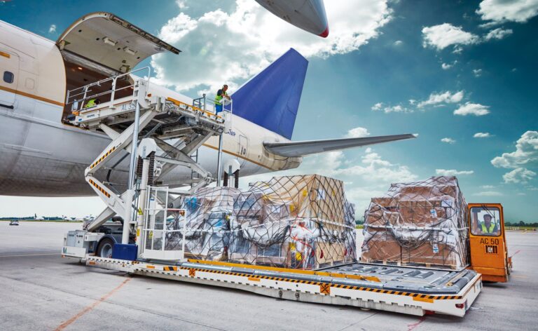 Cargo being loaded onto an airplane on the tarmac, with several packages secured on a pallet and workers operating a lift under a partly cloudy sky.