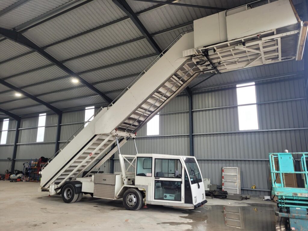 Mobile airplane stairs parked inside a large warehouse with a corrugated metal interior.|A mobile aircraft boarding stair vehicle is parked inside a large warehouse.|Interior of a vehicle with black vinyl seats arranged in two rows. The floor is covered with diamond plate flooring. Large windows provide a view of the outside.|Interior of a control cabin with two black seats