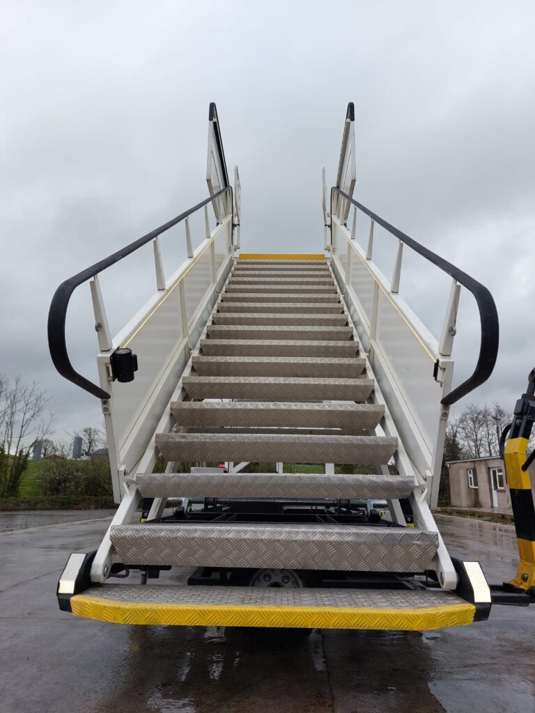 Metal stairs with handrails leading up to an elevated platform on a rainy day. Overcast sky in the background.|Mobile aircraft stairs on a tarmac