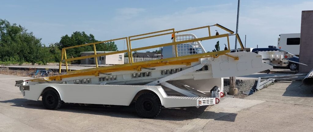 A white trailer with a yellow-railed ramp is parked on a concrete surface near some trees and buildings.|A white airport ground support vehicle with a conveyor belt attachment parked on a concrete surface