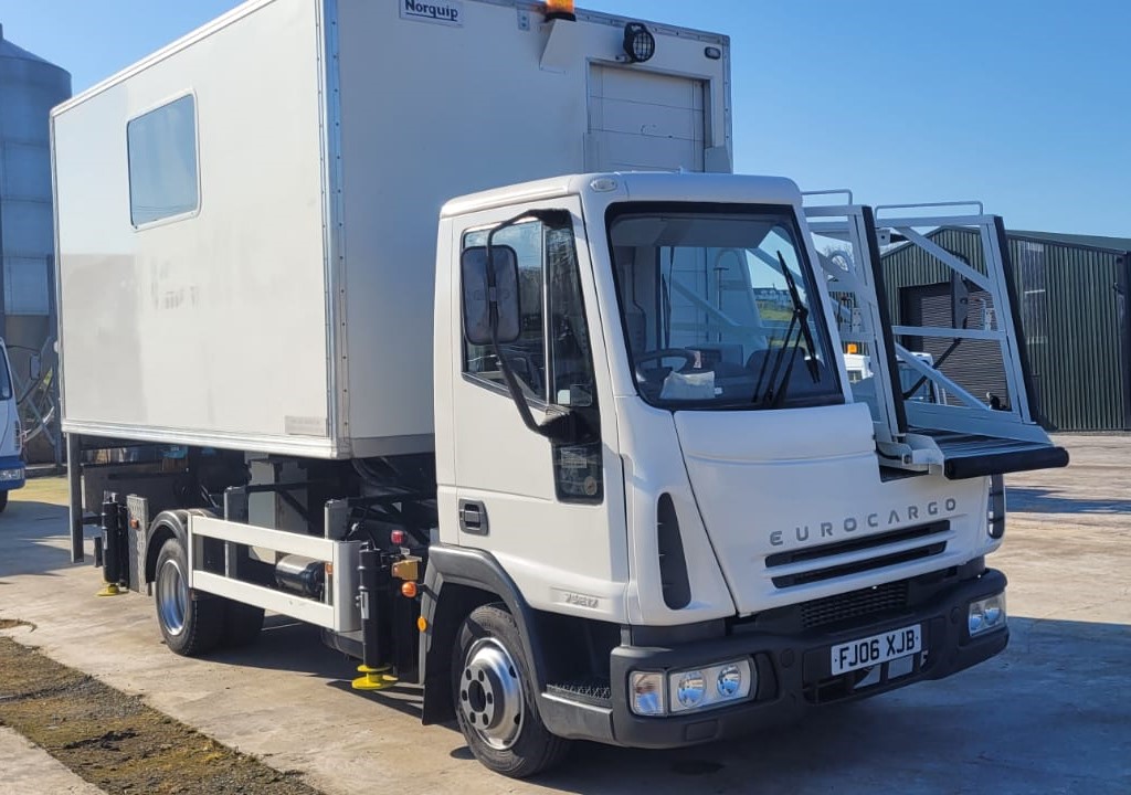 A white Eurocargo truck with a rear compartment is parked on a sunny day.|A white box truck with a liftgate is parked in an open area under a clear blue sky.|