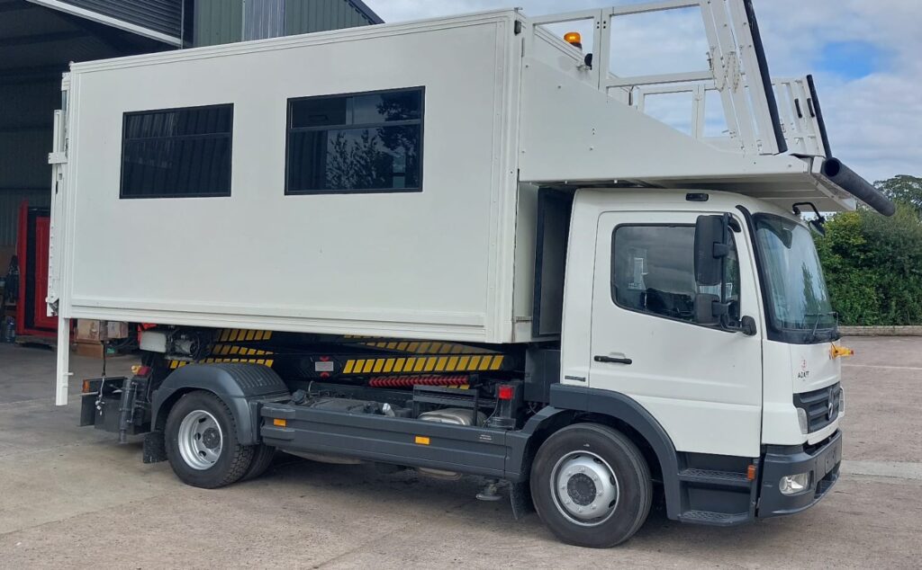 A white mobile unit truck with a large box-like structure and windows is parked in an industrial area.|A white mobile platform truck with an extended cabin stands on a concrete surface near a building.|White mobile office truck parked on a concrete surface with a door and window on the side.|