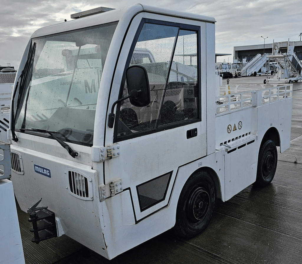 White airport baggage tug on a wet tarmac near boarding ramps.|Interior view of a vehicle cabin with two black leather seats